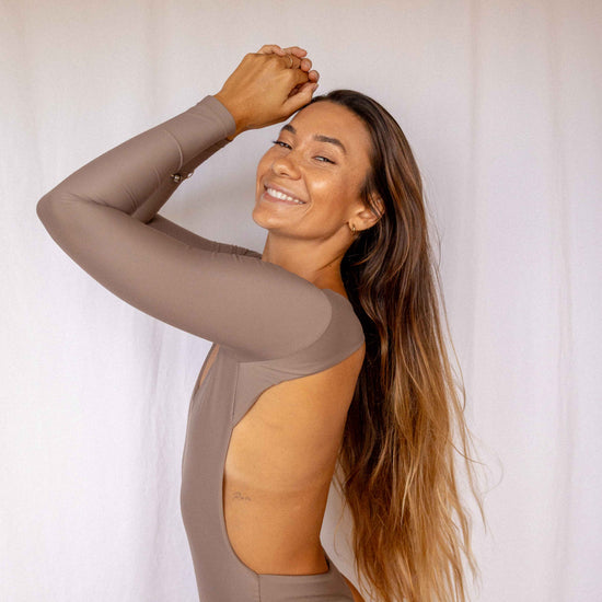 Nia Long-Sleeve Surf Suit in Taupe, smiling side profile view showing open back cutout detail and long-sleeve construction by Antidote The Label