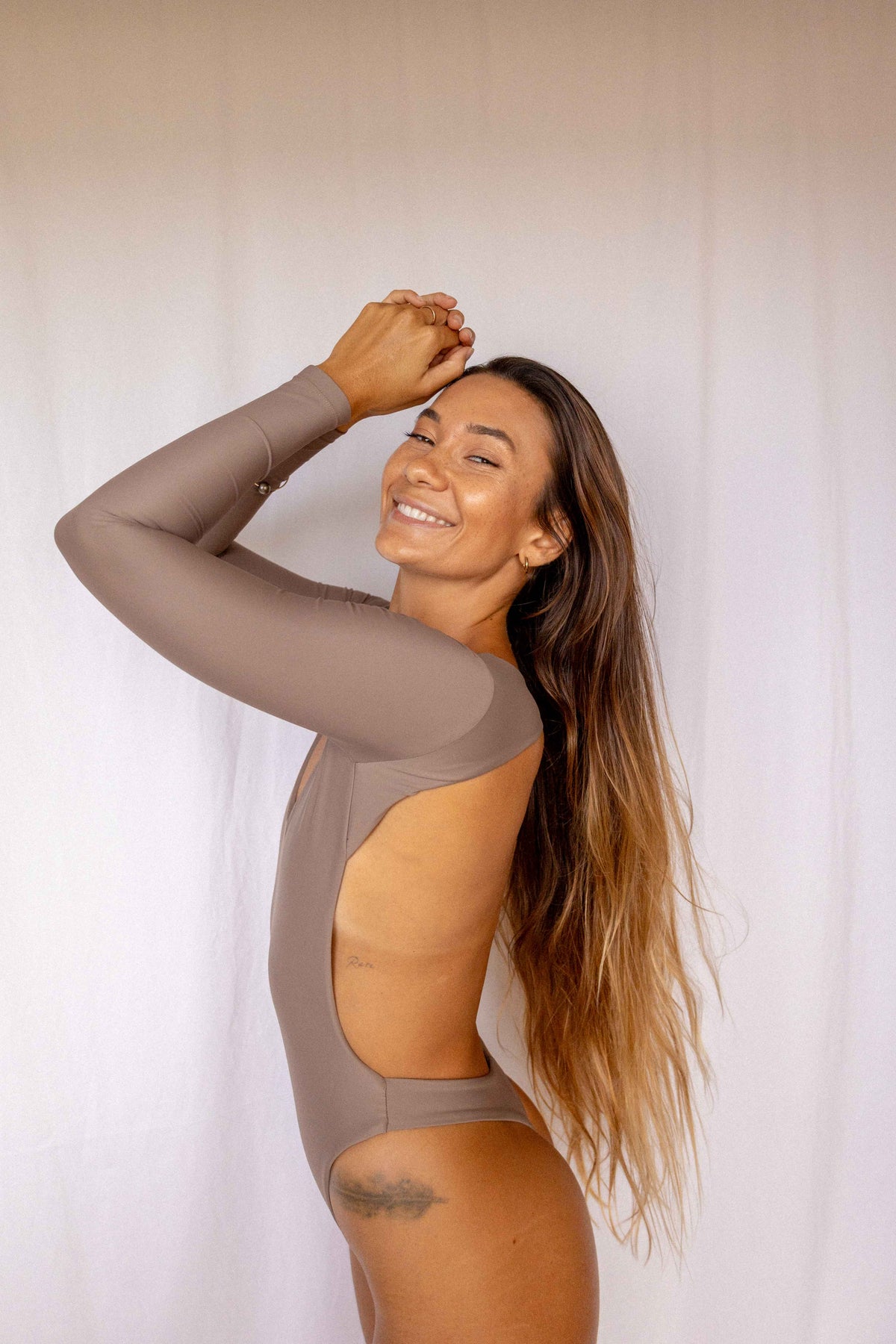 Nia Long-Sleeve Surf Suit in Taupe, smiling side profile view showing open back cutout detail and long-sleeve construction by Antidote The Label