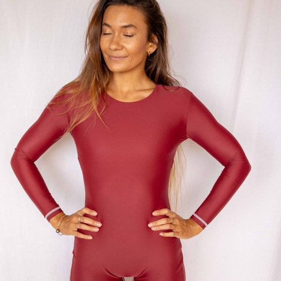 Anou Long-Sleeve Surf Suit in Deep Merlot, smiling full front view with hands on hips showing round neckline, long-sleeve fit, and high-cut short silhouette by Antidote The Label