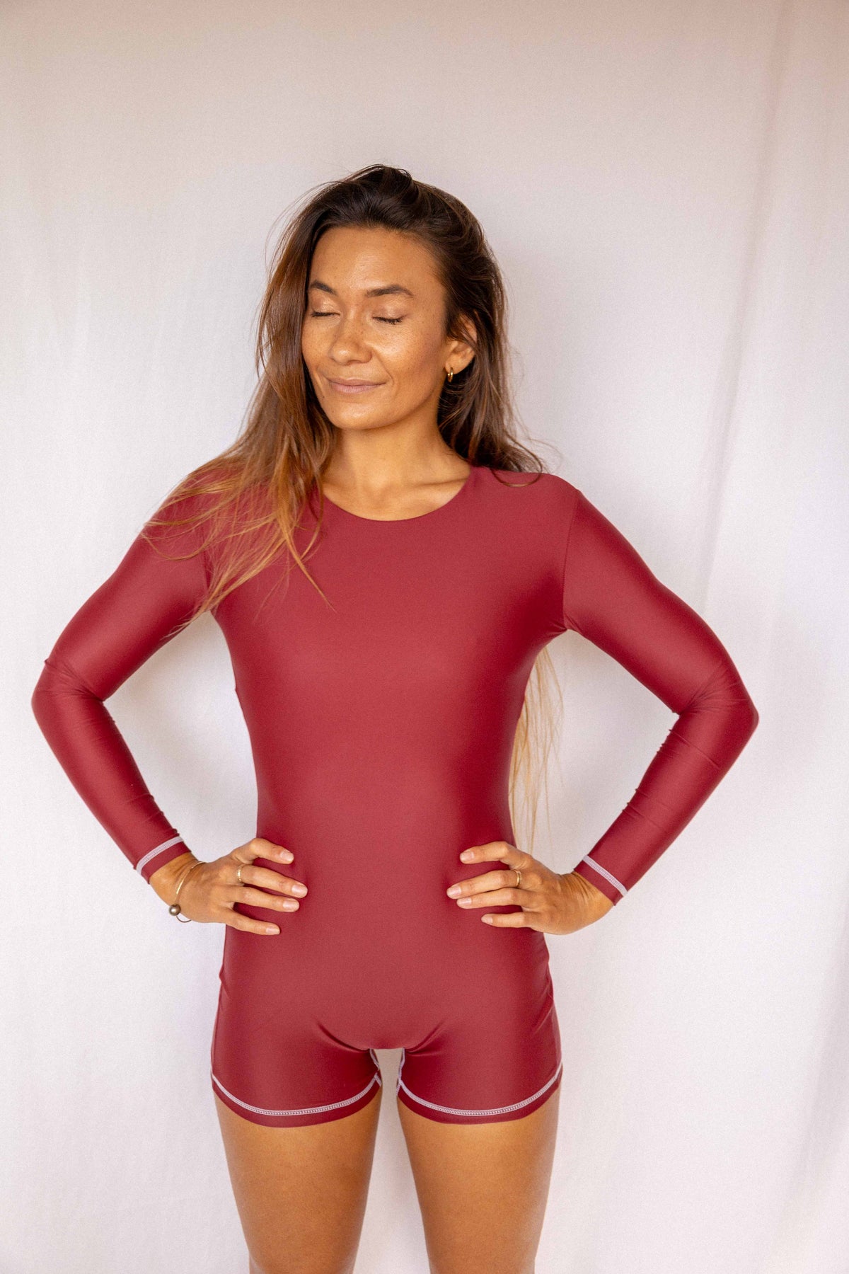 Anou Long-Sleeve Surf Suit in Deep Merlot, smiling full front view with hands on hips showing round neckline, long-sleeve fit, and high-cut short silhouette by Antidote The Label
