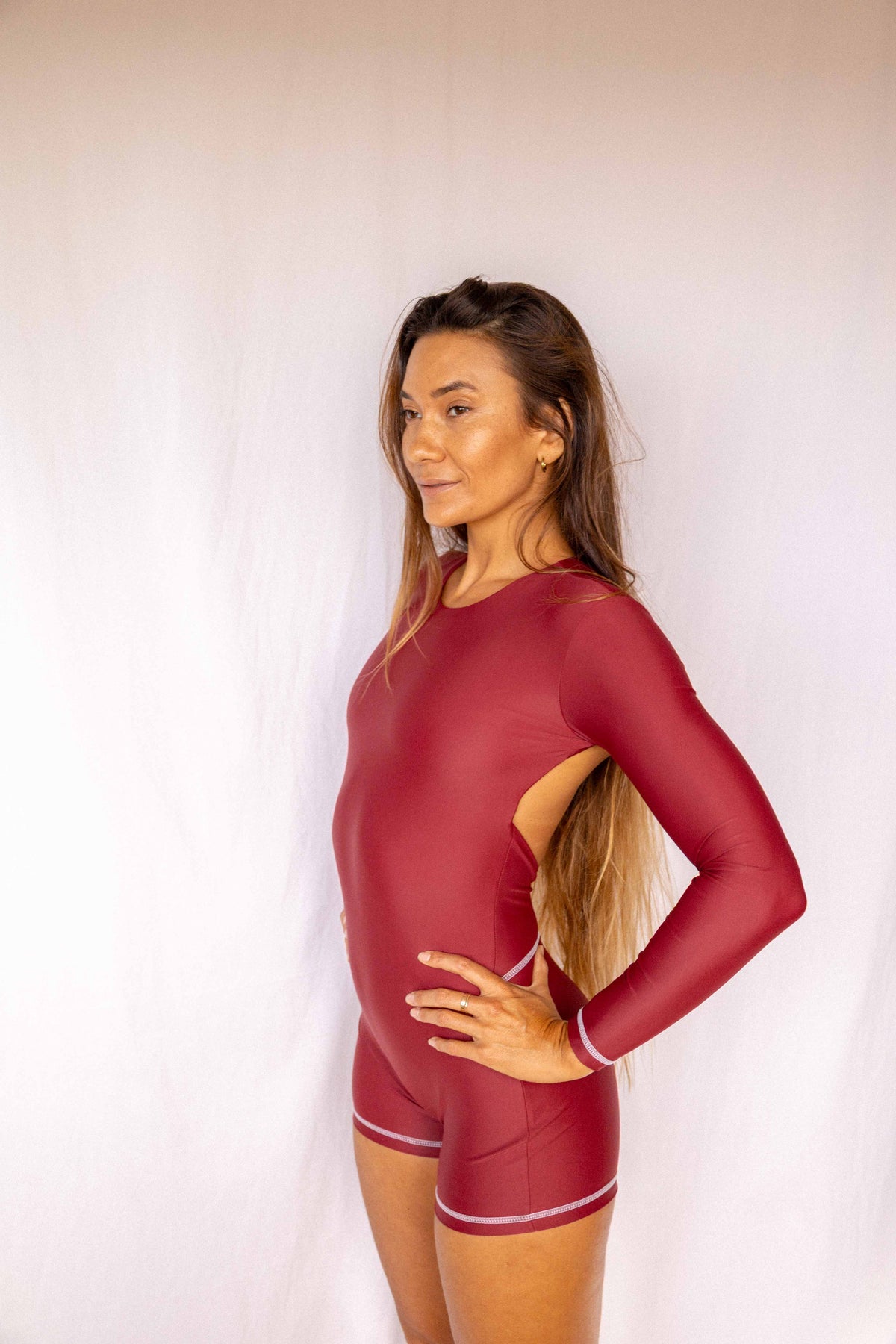 Anou Long-Sleeve Surf Suit in Deep Merlot, side profile showing open back cutout and streamlined long-sleeve one-piece silhouette