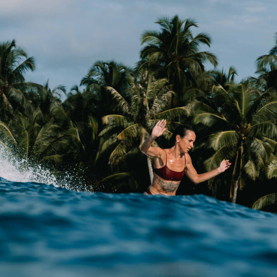 Ona Surf Top in Deep Merlot worn surfing in ocean waves with tropical backdrop, women's performance surf top by Antidote The Label
