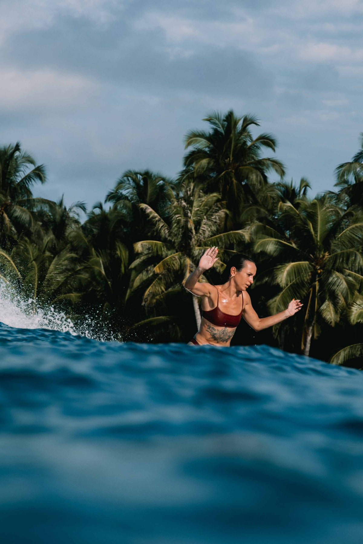 Ona Surf Top in Deep Merlot worn surfing in ocean waves with tropical backdrop, women's performance surf top by Antidote The Label